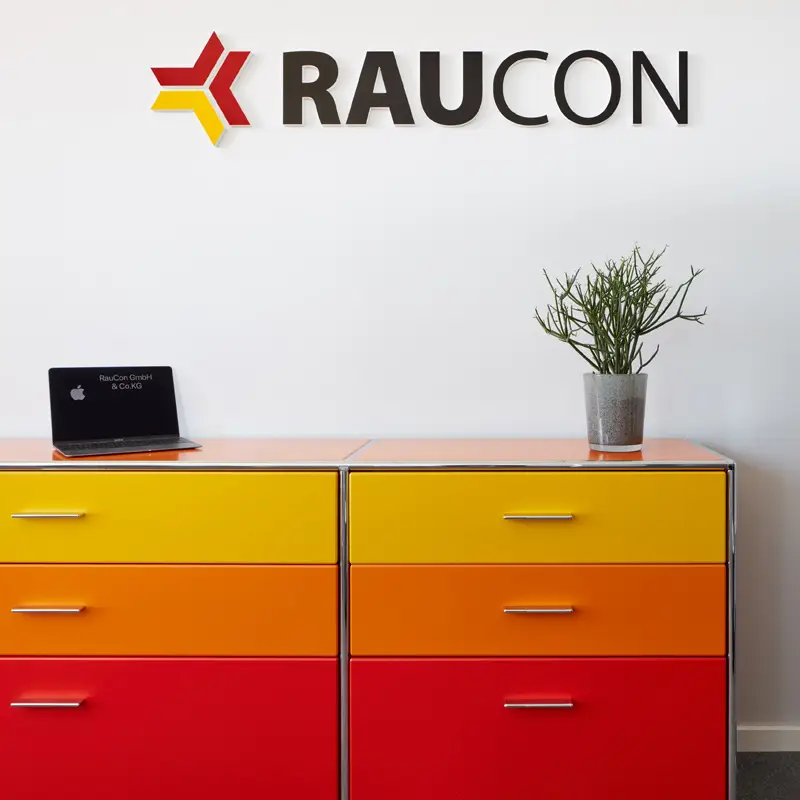 Laptop und Pflanze auf bunter Kommode vor weißer Wand mit RAUCON-Logo.