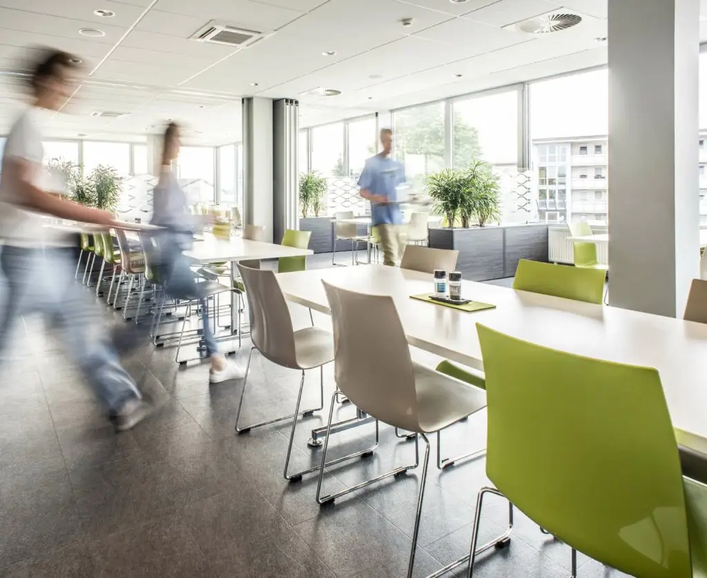Heller, moderner Pausenraum mit grünen und weißen Stühlen, großen Fenstern und Pflanzen im Hintergrund.
