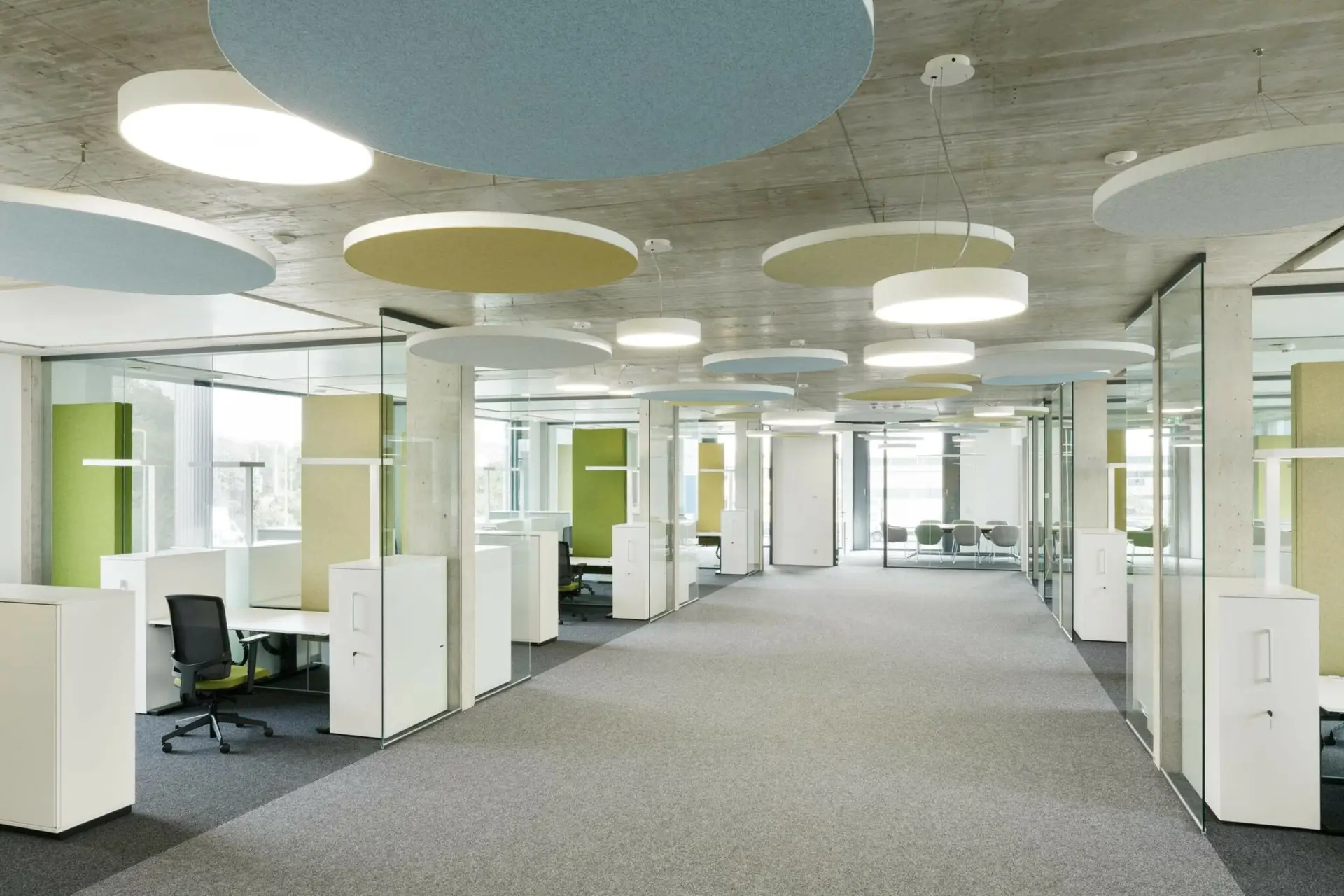 Moderner Büroraum mit Glaswänden, Arbeitsplatznischen, bunten Deckenelementen, Teppichboden.