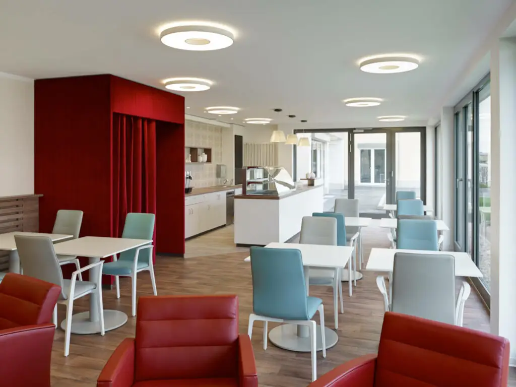 Moderner Raum mit roten, grauen und blauen Stühlen, weißen Tischen und großen Fenstern.