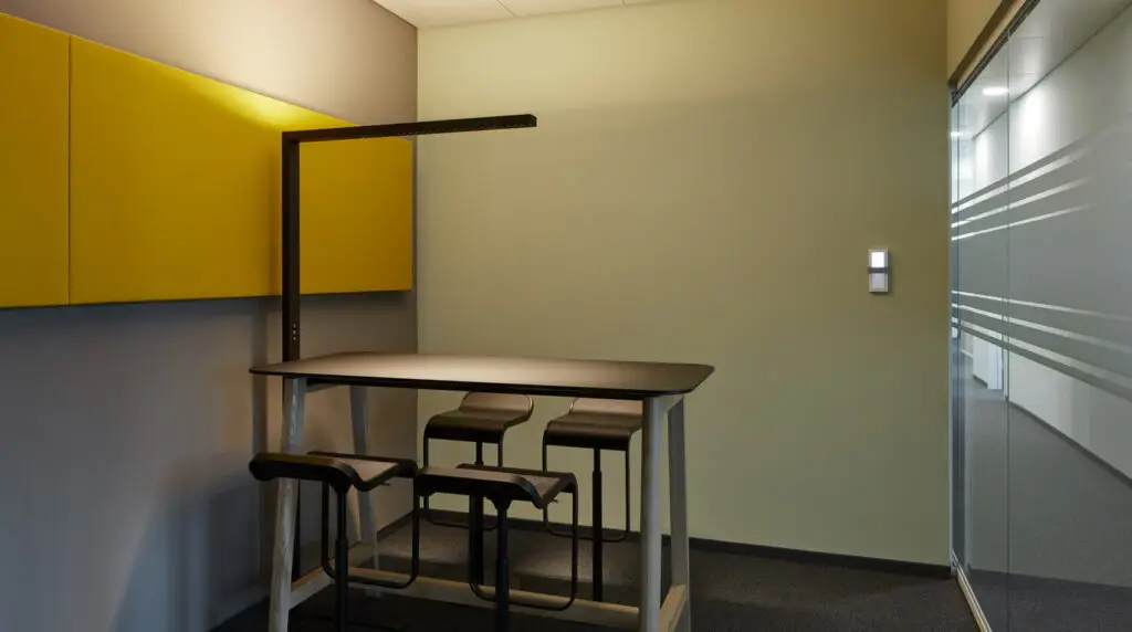 Moderner Raum mit hoher schwarzer Tischfläche, vier Stühlen, gelben Akustikpaneelen und Glastrennwand.