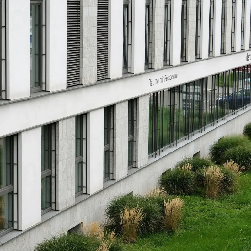 Modernes Bürogebäude mit großen Fenstern, Steinverkleidung und gepflegter Grünfläche davor.